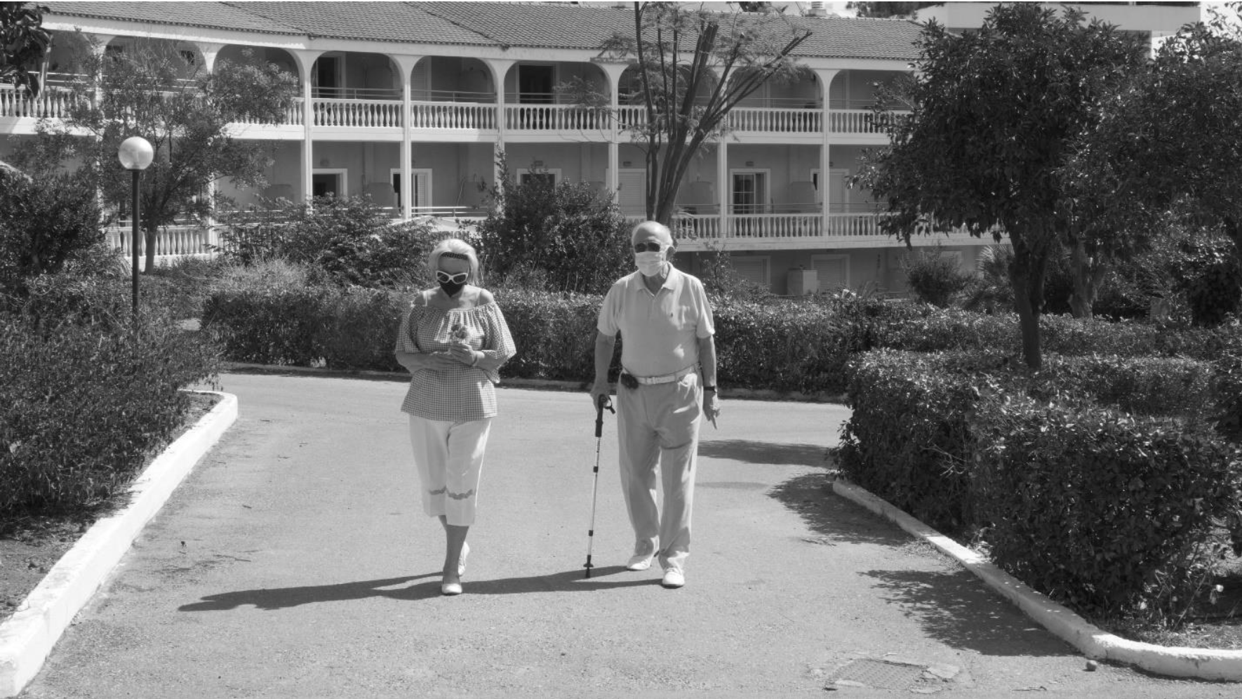 Ασπρόμαυρη εικόνα δύο ηλικιωμένων, ενός άντρα και μίας γυναίκας, να περπατάνε σε ανοιχτό χώρο φορώντας μάσκες για την προστασία από τον ιό